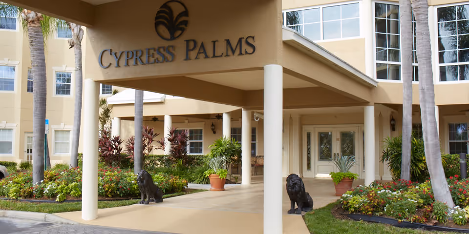 Entrance of Cypress Palms facility with a covered driveway supported by white columns, two black lion statues on either side, surrounded by landscaped greenery and palm trees, and a beige building in the background.