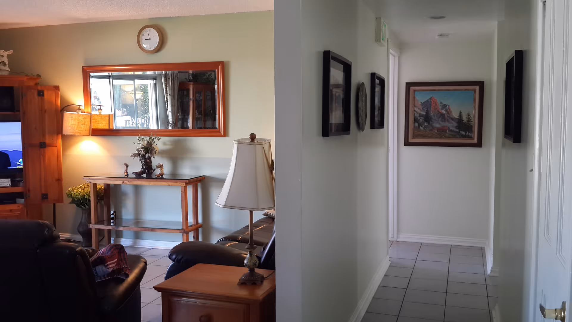 Interior view showing a cozy living room area with leather chairs, a wooden side table with a lamp, a wooden console table with decorative items, a wall clock above a large mirror, and a hallway with framed pictures and a painting at the end.
