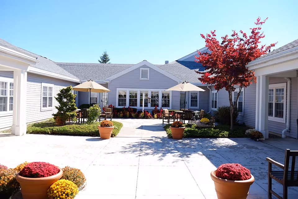 Outdoor courtyard area of a senior living facility with potted flowers, green shrubs, and trees. There are patio tables with umbrellas and chairs arranged for seating. The building surrounding the courtyard is light gray with white trim and multiple windows. The sky is clear and blue.