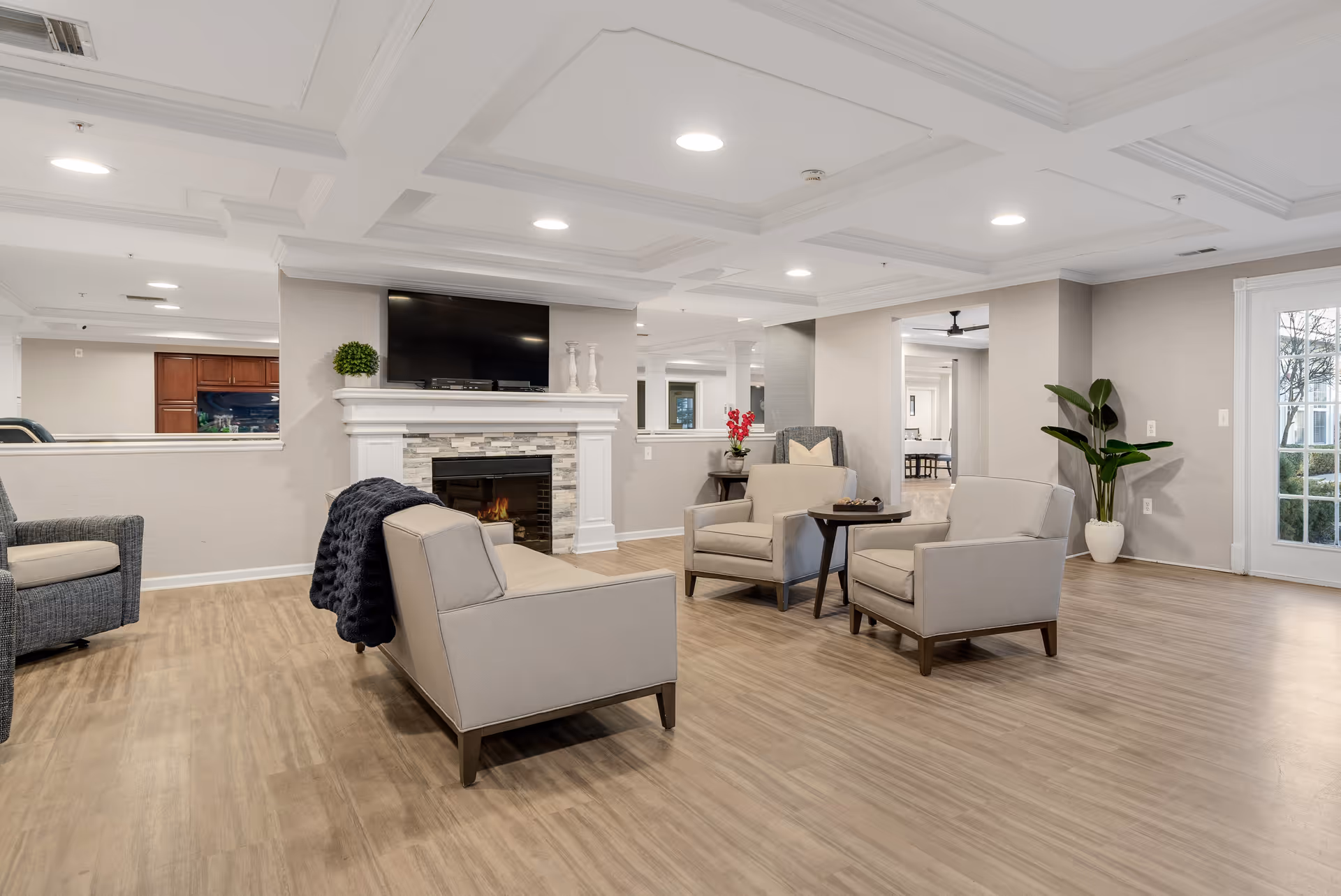 A spacious and well-lit living room area with light wood flooring, beige walls, and a white coffered ceiling with recessed lighting. The room features a white fireplace with a mounted flat-screen TV above it, two beige armchairs, a beige loveseat with a dark throw blanket, a small round wooden table, a gray armchair, and a potted plant near a glass door leading outside.