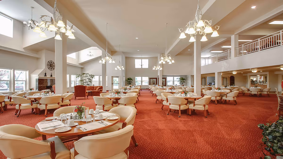 Spacious dining room with round tables set for meals, cream chairs, red carpet, chandeliers, and a mezzanine level.