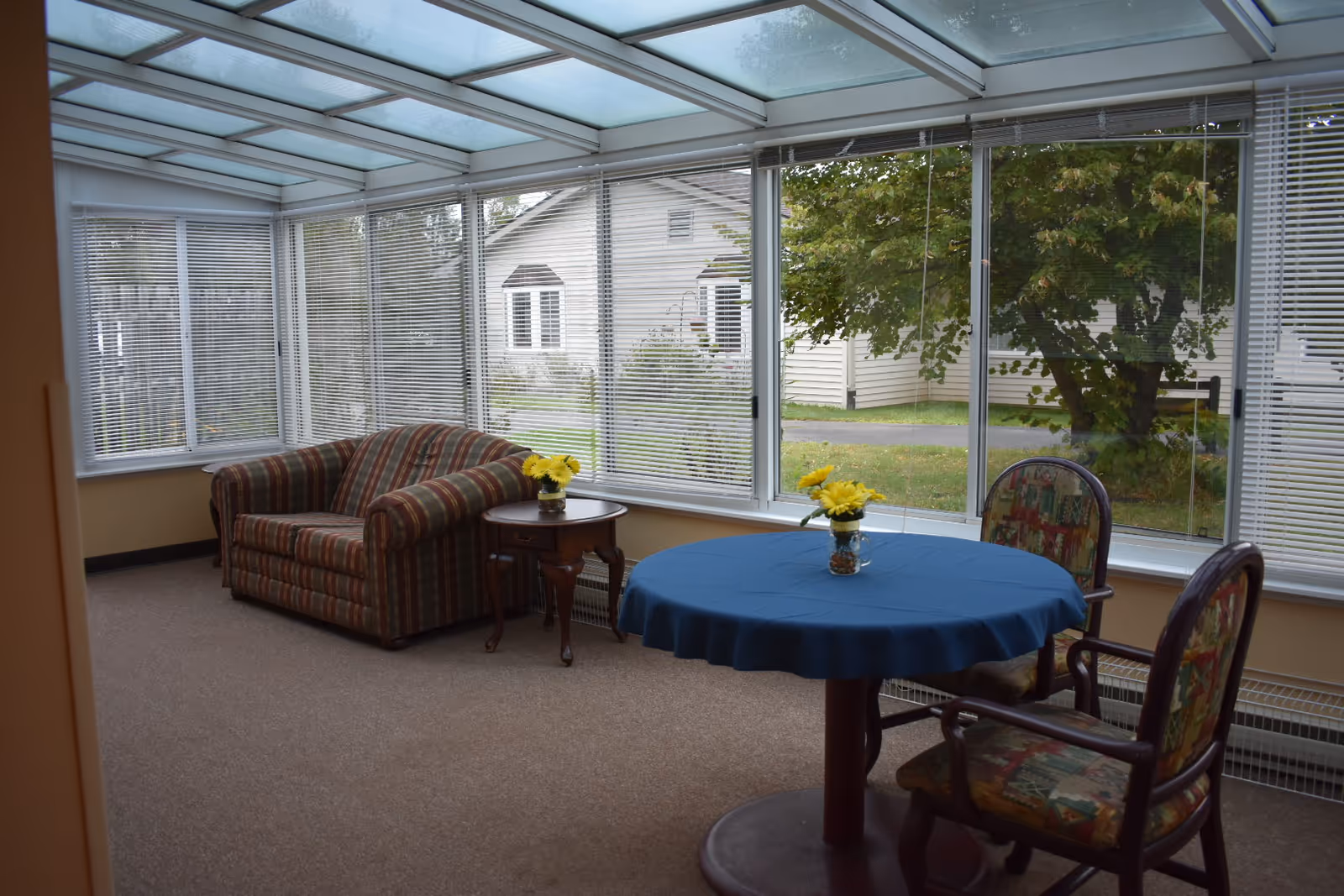 A sunroom with large windows and a glass ceiling, featuring a striped loveseat, a small wooden side table with a vase of yellow flowers, and a round table covered with a blue tablecloth surrounded by two upholstered chairs, also with a vase of yellow flowers on the table. Outside the windows, a house and trees are visible.