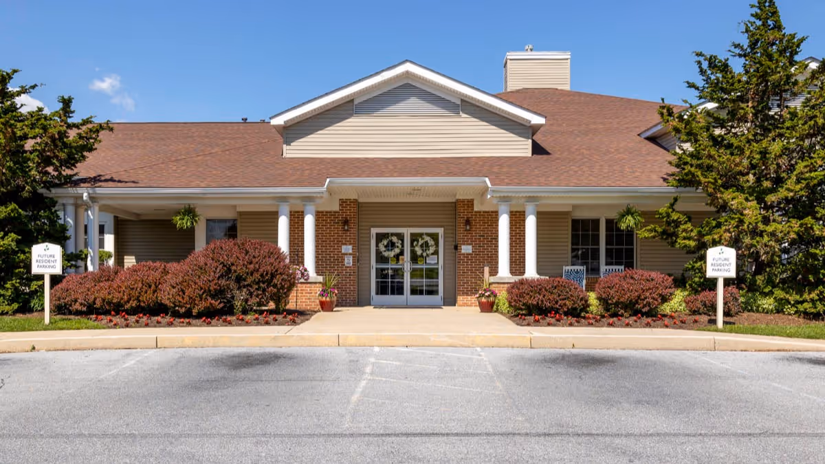 Front entrance of a single-story senior living building with white columns, landscaped shrubs, and empty parking spaces.