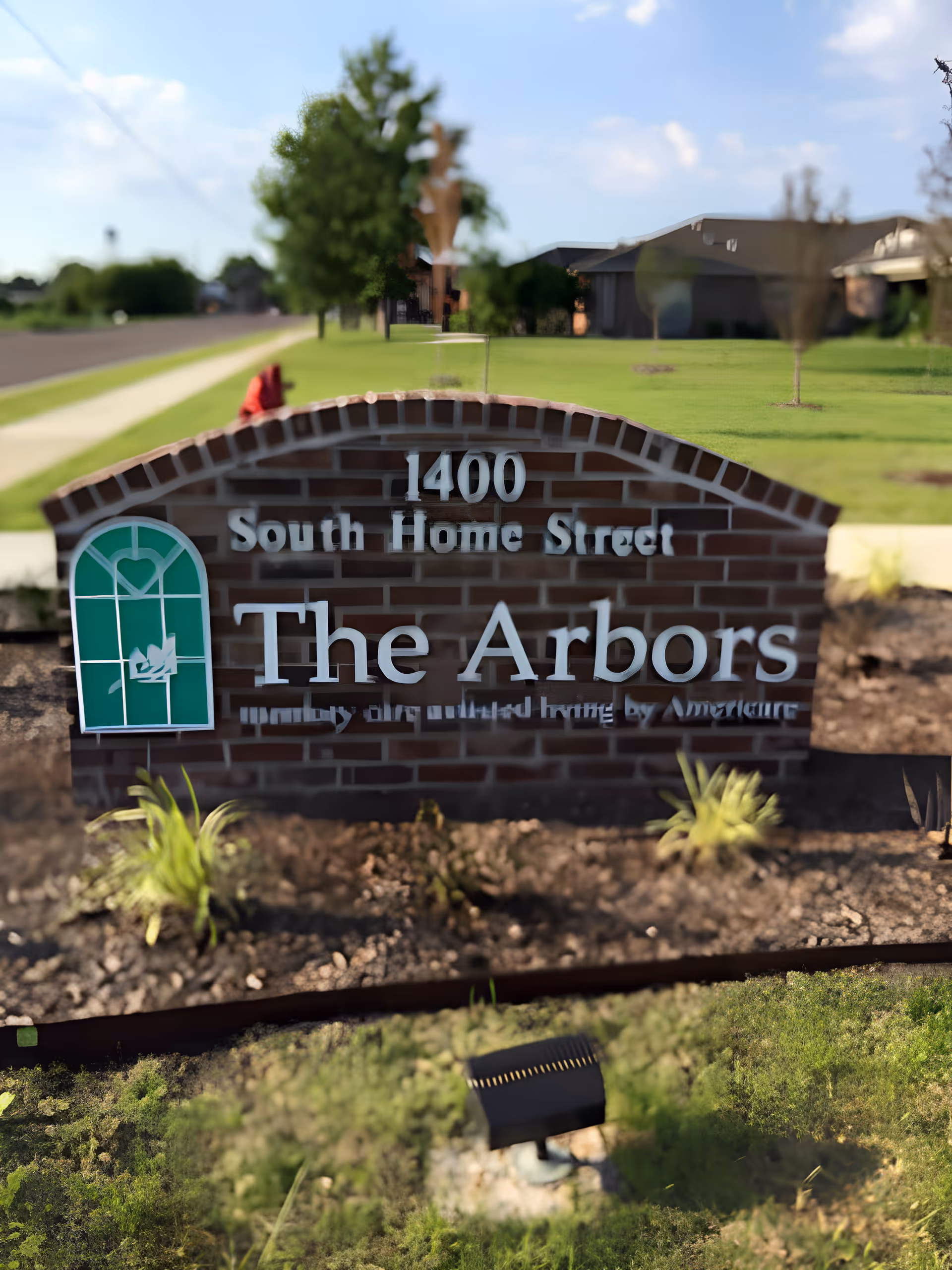 A brick sign outside a facility with the address 1400 South Home Street. The sign reads 'The Arbors' with a green window-like logo featuring a heart and a bird. The background shows a grassy area, a sidewalk, and some buildings under a partly cloudy sky.