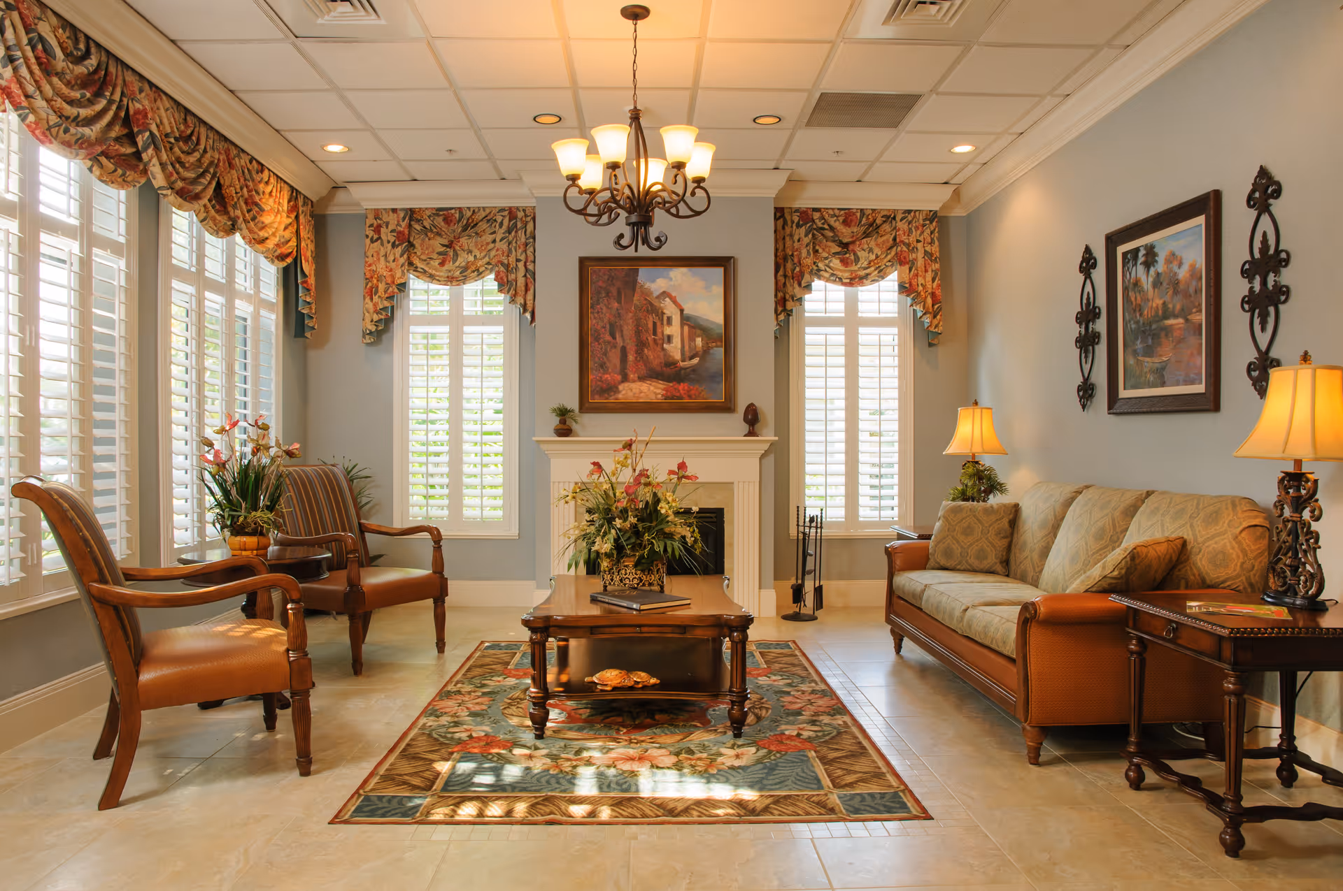 A warmly lit living room with a beige sofa and two wooden armchairs with leather seats. A wooden coffee table with a floral arrangement sits on a patterned area rug in front of a white fireplace. The room has large windows with white shutters and floral valances. Two table lamps with beige shades and framed artwork adorn the walls.