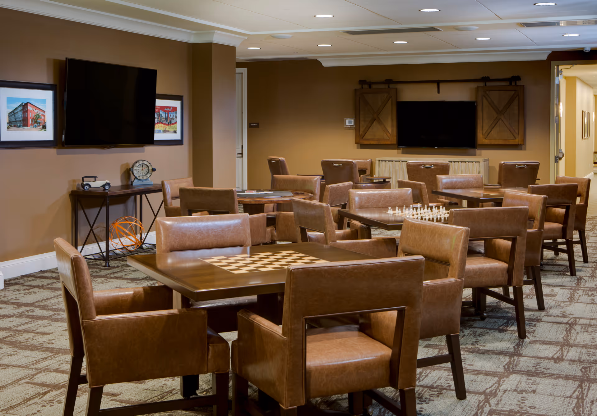 A cozy common area with multiple tables and brown leather chairs arranged for games like chess and checkers. The room has warm brown walls, a patterned carpet, two flat-screen TVs mounted on the walls, and framed artwork. A small console table with decorative items is against one wall.