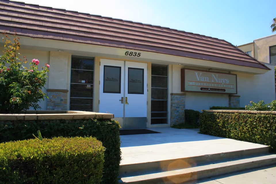 Front entrance of a healthcare facility with double white doors, the address number 6835 above the doors, and a sign on the right reading 'Van Nuys Healthcare Center'. There are bushes and flowers on either side of the entrance and a clear blue sky above.