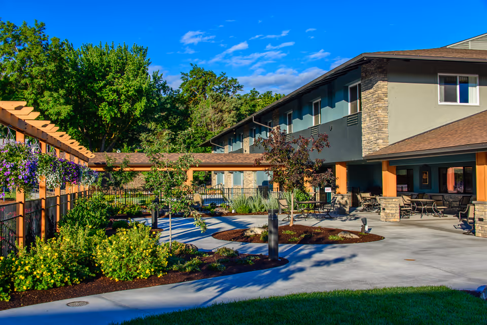 Courtyard of a senior living facility with paved walkways, landscaped gardens, a pergola with hanging flowers, and outdoor seating by a two-story building.