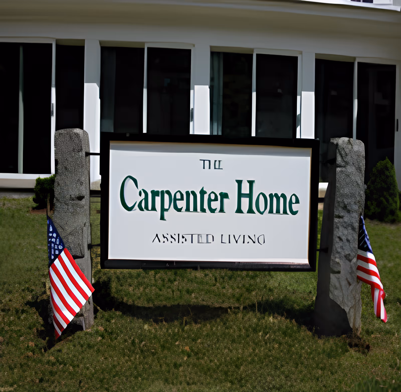 A sign for The Carpenter Home assisted living facility mounted between two stone posts with small American flags placed at the base of each post, in front of a building with large windows.