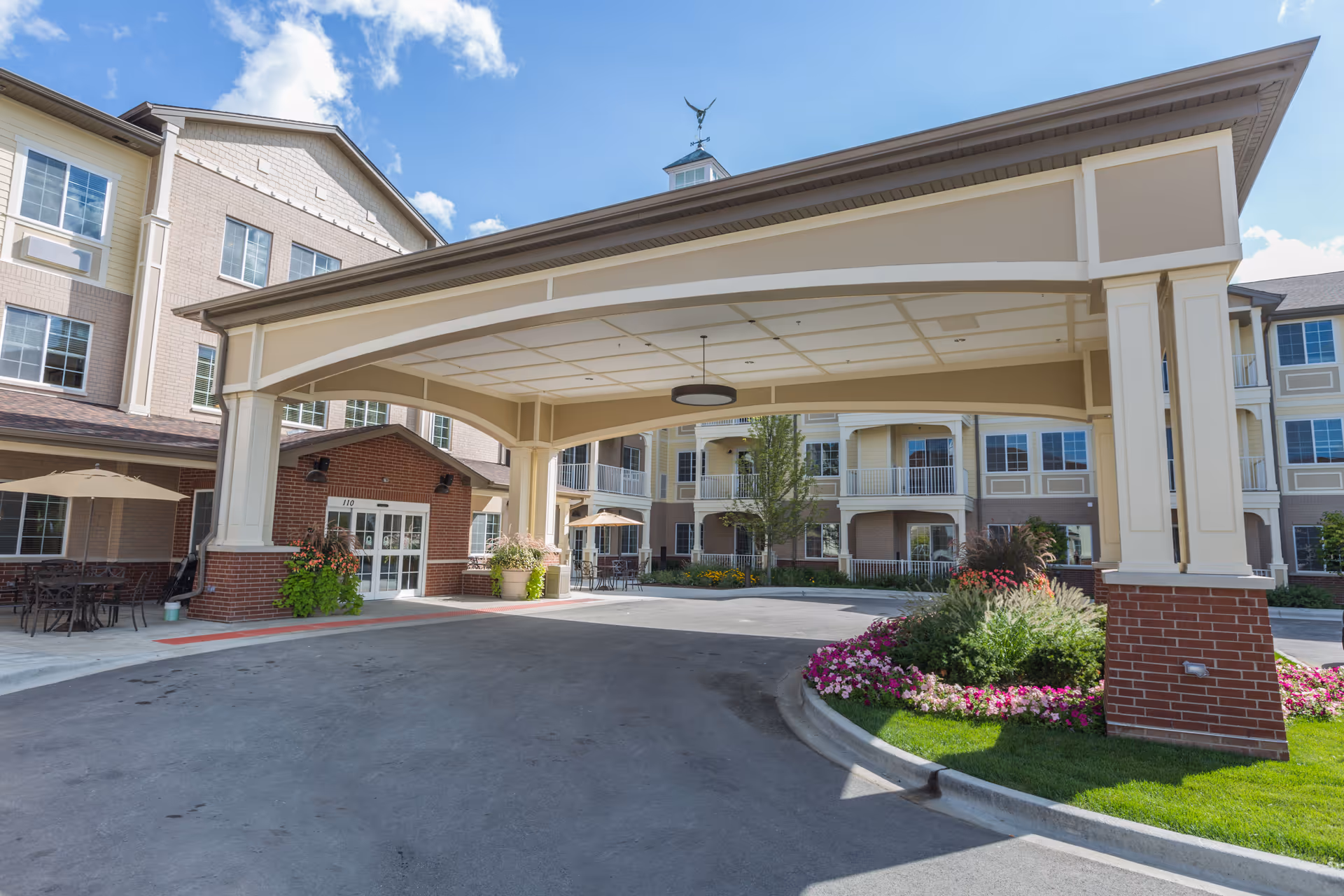 Entrance area of Westbrook Senior Living facility showing a covered drop-off zone with a large beige canopy supported by columns. The building has multiple floors with balconies and large windows. There are flower beds with colorful flowers and greenery near the entrance, and outdoor seating with tables and umbrellas is visible on the left side.