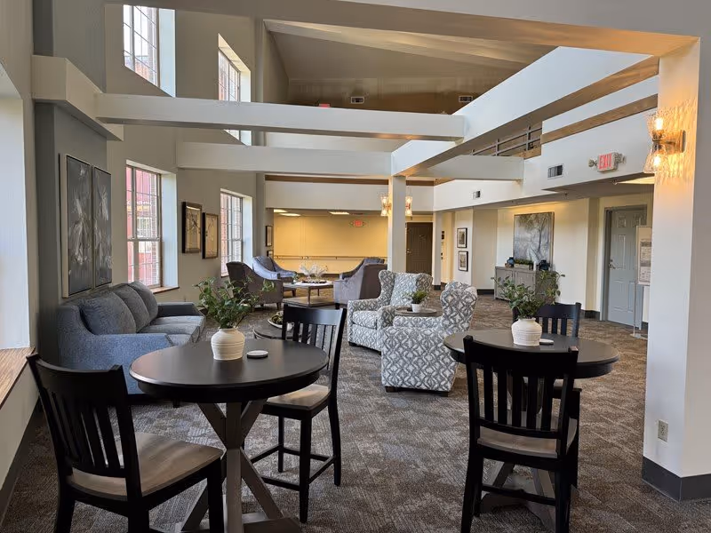 A spacious and well-lit common area in a retirement community featuring multiple seating arrangements including round tables with chairs, cushioned armchairs, and sofas. The room has large windows allowing natural light, modern light fixtures on the walls, and neutral-toned carpet flooring. Decorative plants and framed artwork adorn the space, creating a welcoming and comfortable atmosphere.