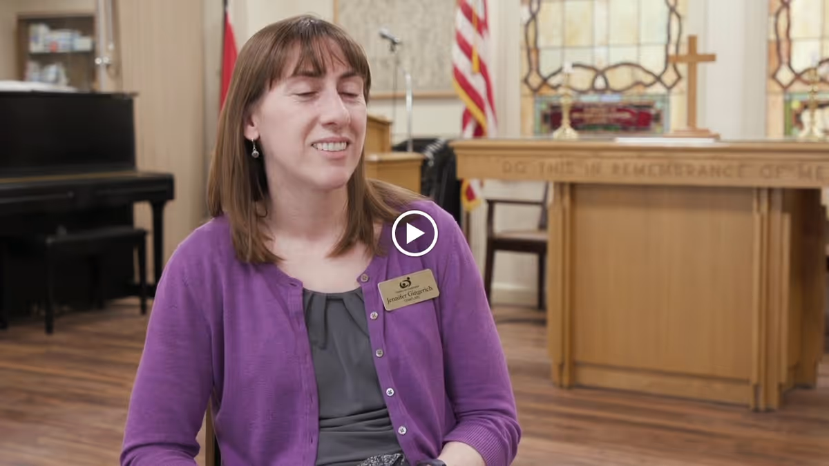 A woman wearing a purple cardigan and a name tag sits in a room with wooden floors, a piano, and a wooden altar with a cross and stained glass windows in the background.