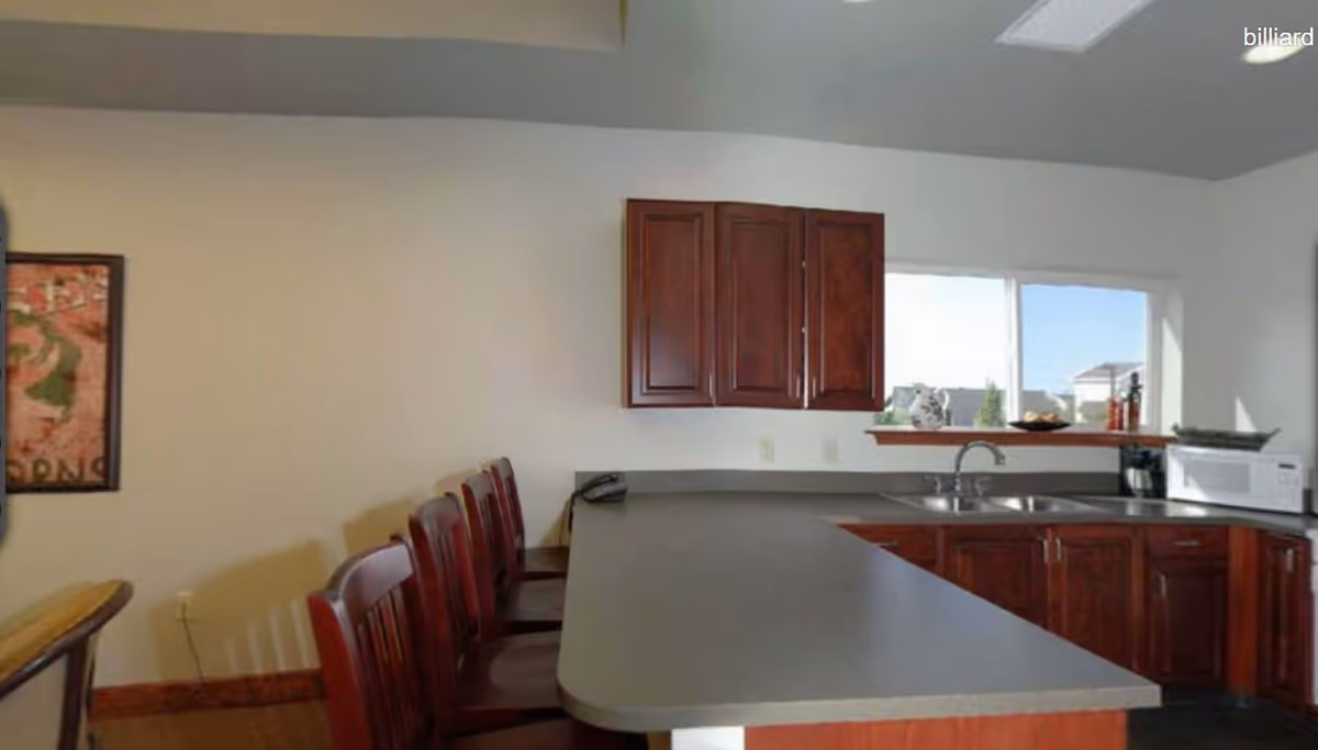 Interior view of a kitchen area with a long countertop and several wooden chairs lined up along one side. The kitchen has dark wood cabinets, a double sink under a window, a microwave, and some kitchen items on the counter. A framed picture is partially visible on the left wall.
