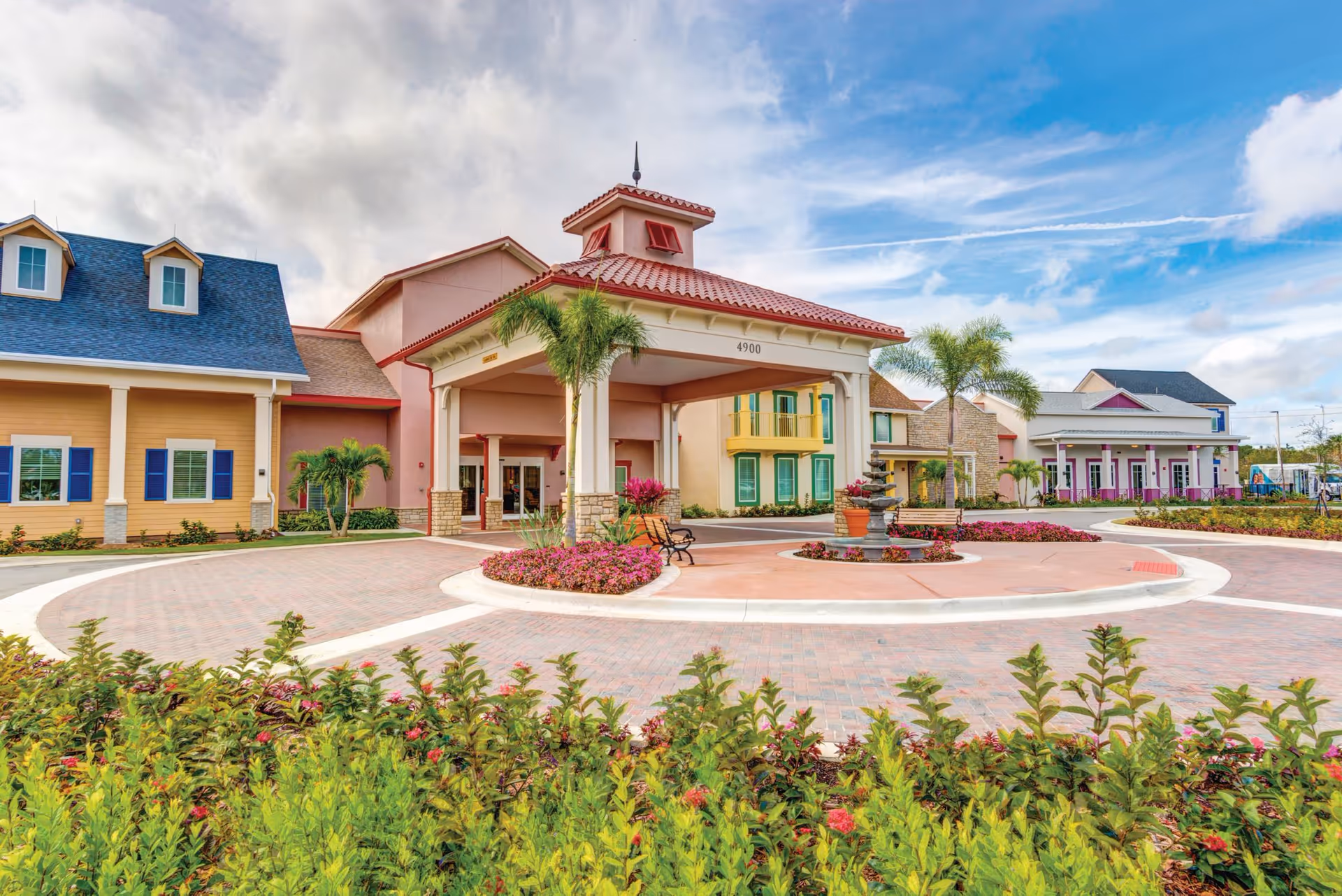 Sunny senior living campus with a pink-tan building, circular drive, palm trees, and a fountain.