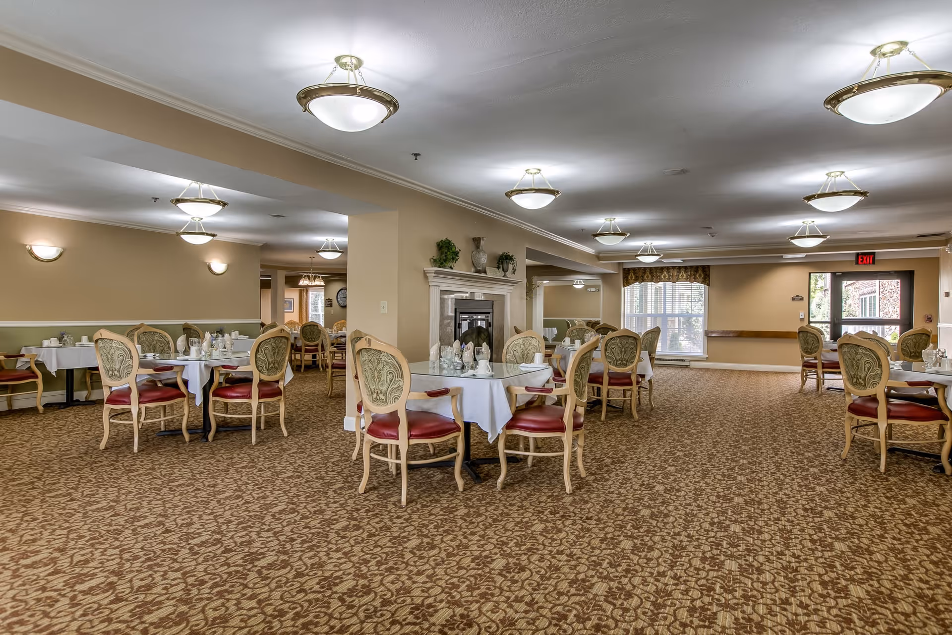 A spacious dining room in a retirement community with multiple round tables covered with white tablecloths, each surrounded by ornate chairs with red cushions. The room has beige walls, patterned carpet, ceiling lights, and large windows letting in natural light. There is a fireplace in the center of the room and an exit door at the far end.