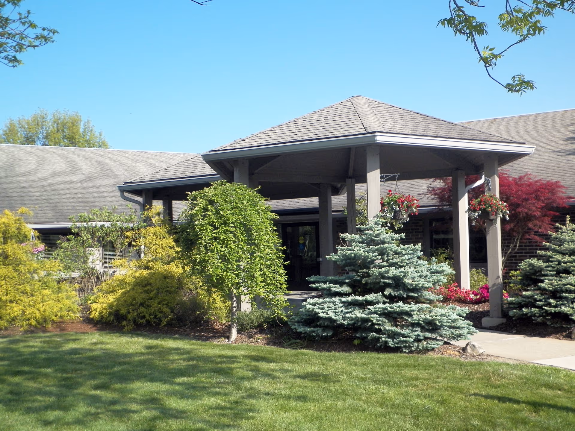 Entrance to a single-story building with a covered porch supported by columns, surrounded by well-maintained landscaping including various shrubs, small trees, and hanging flower baskets under a clear blue sky.