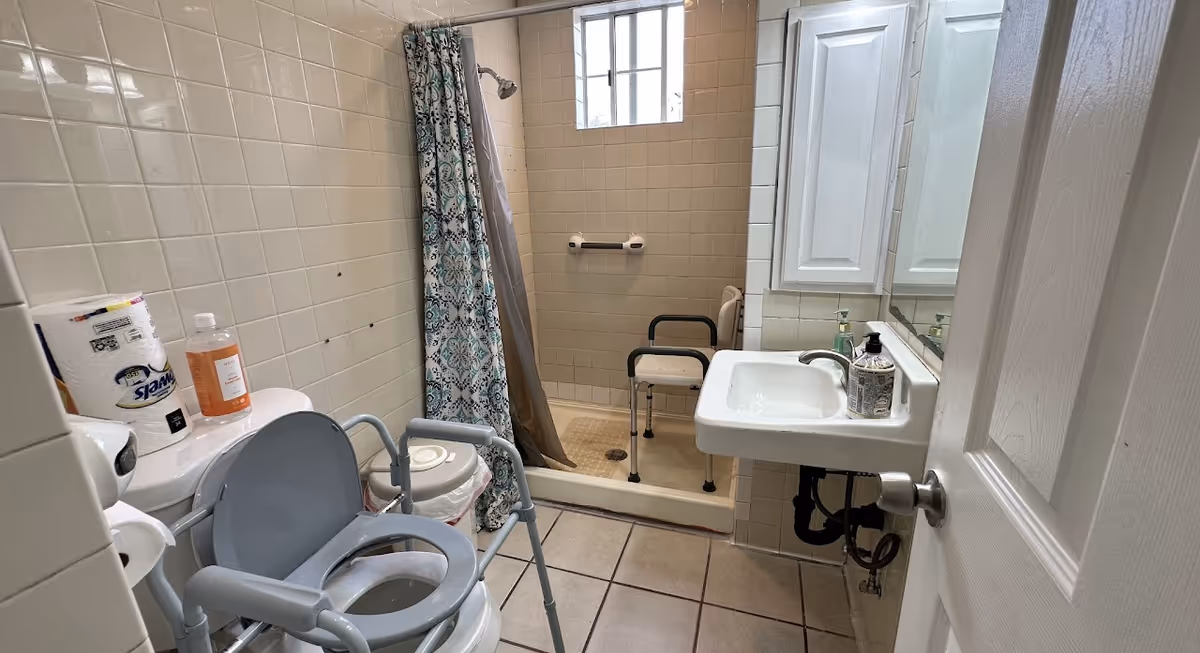 A small bathroom with beige tiled walls and floor. It features a toilet with a raised seat and armrests, a roll of paper towels and a bottle of soap on top of the toilet tank. There is a shower area with a patterned shower curtain and a shower chair inside. A white sink with a soap dispenser is mounted on the wall next to a mirrored cabinet. A window above the shower allows natural light into the room.