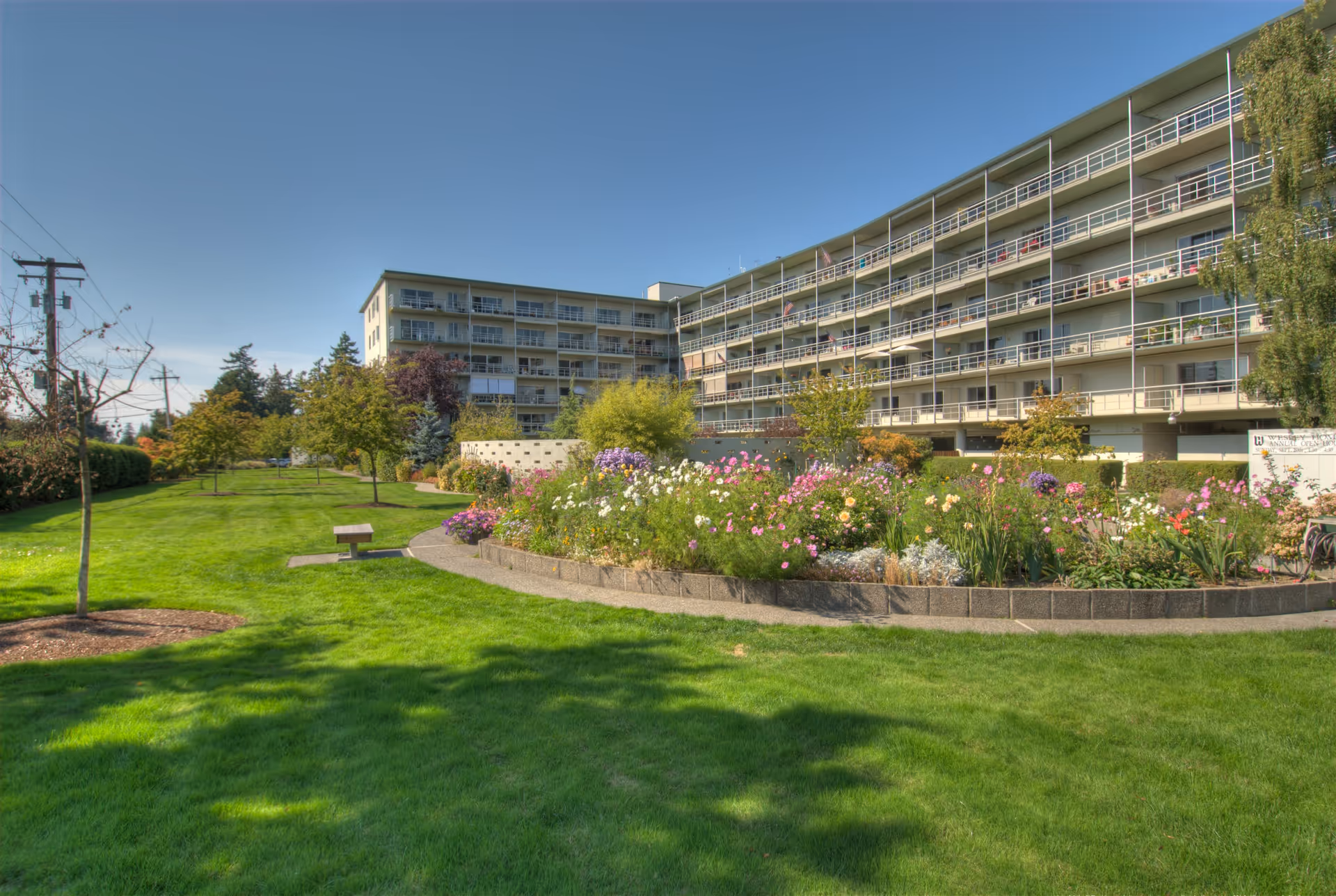 A large multi-story senior living facility building with balconies overlooking a well-maintained garden area featuring colorful flowers, green grass, trees, and a walking path under a clear blue sky.