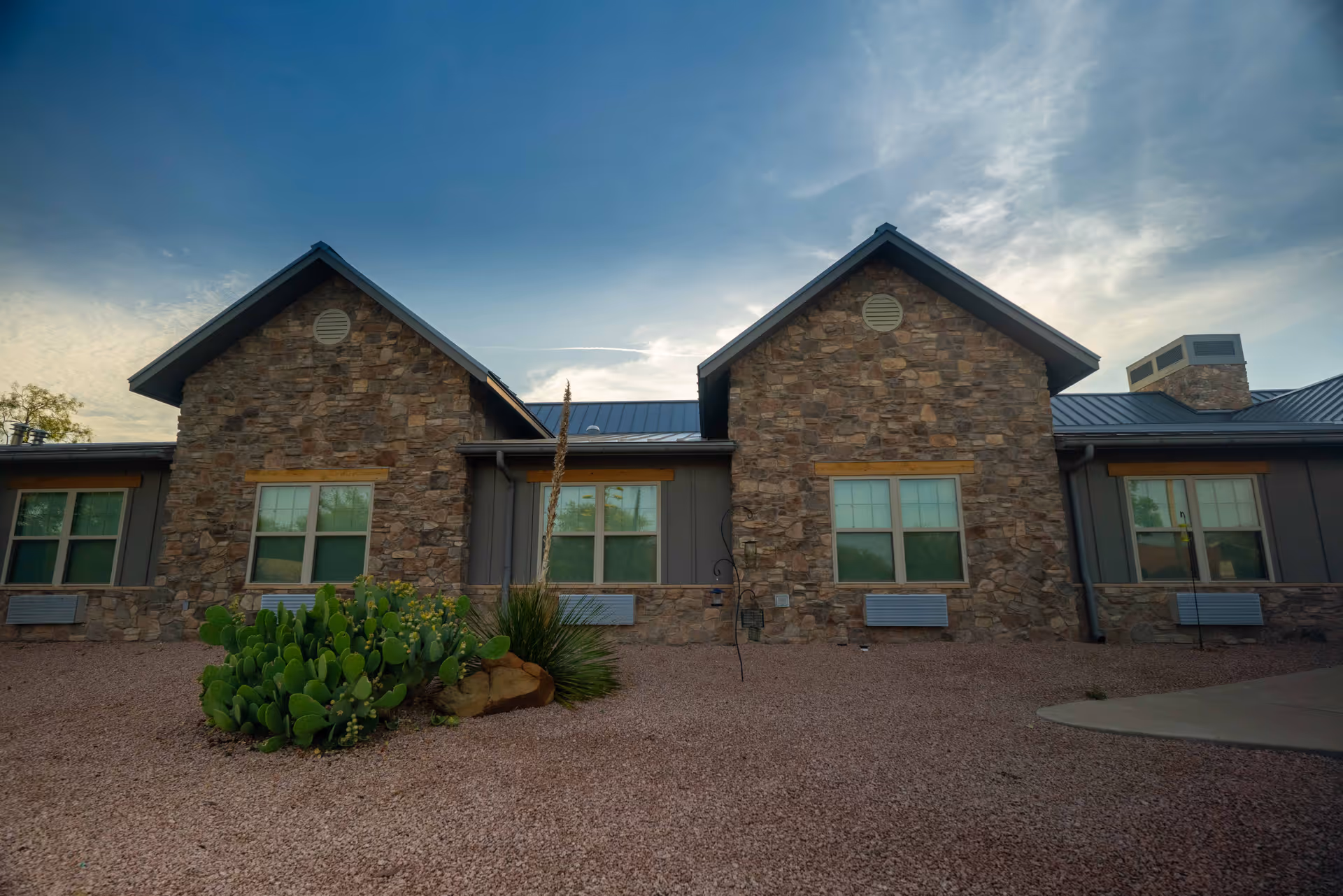 Stone-faced single-story building with gabled roofs, multiple windows, and desert landscaping with cacti under a blue sky.