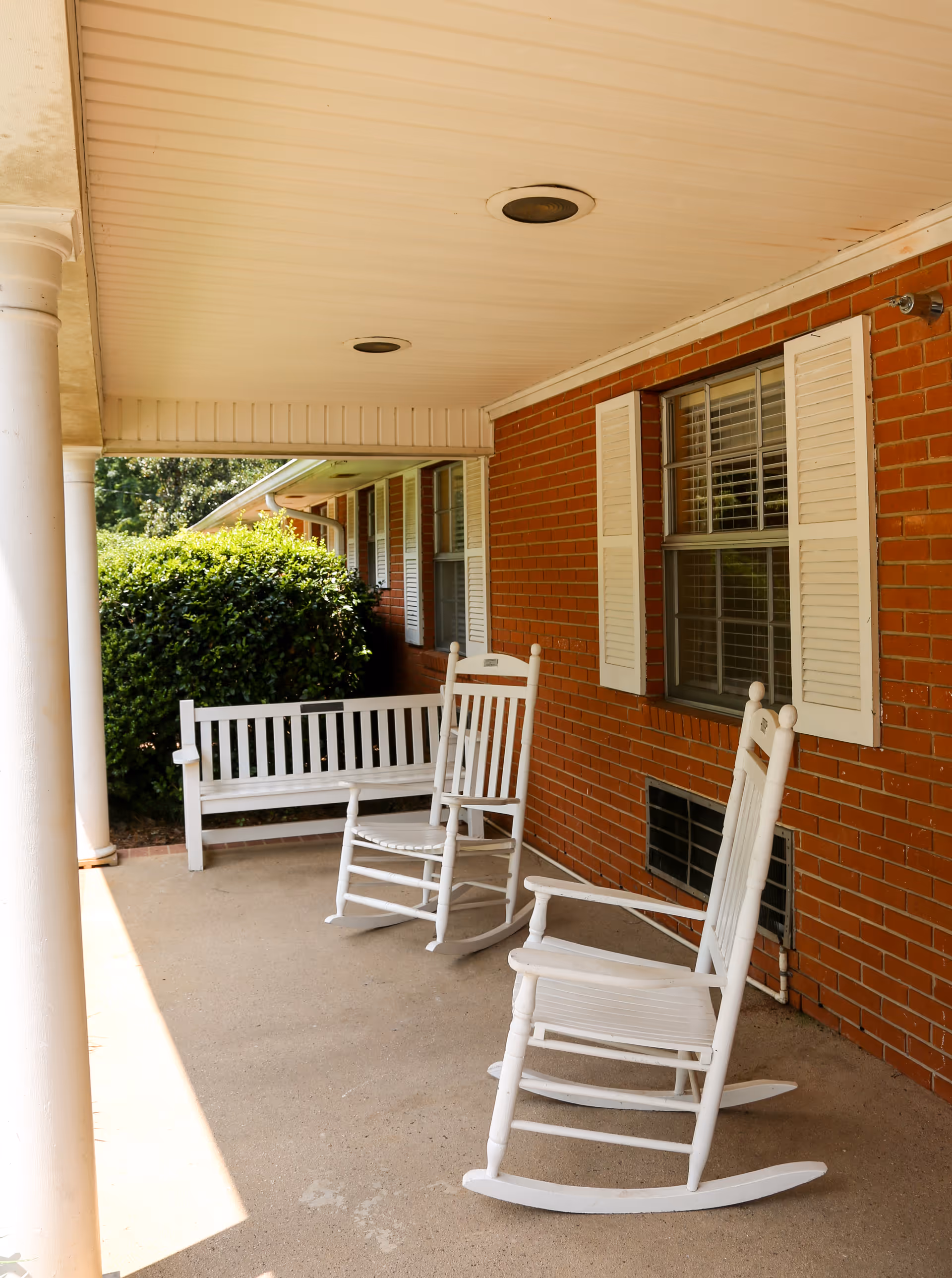 Covered brick front porch with white rocking chairs and a bench beneath a roofed overhang.