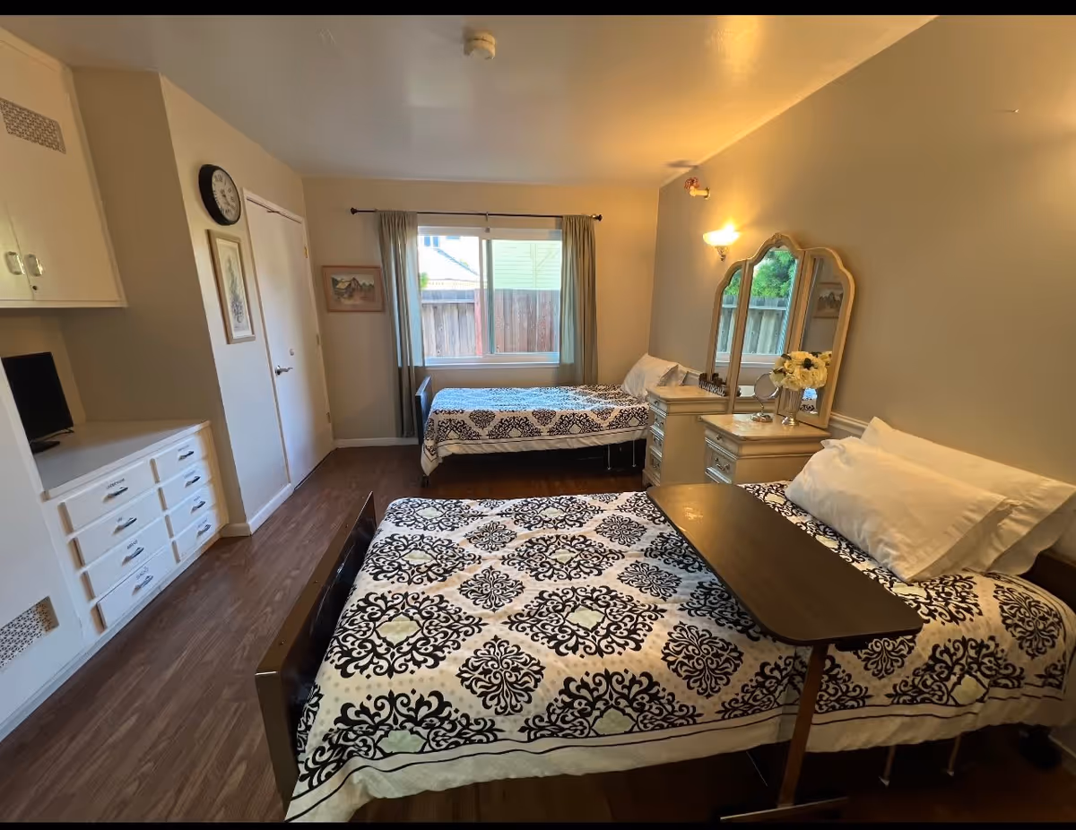 A bedroom with two single beds covered in patterned bedspreads. Each bed has white pillows, and one bed has a small overbed table. There is a dresser with a three-panel mirror and a vase of flowers between the beds. A window with curtains is on the far wall, and a clock and framed pictures hang on the walls. The floor is wood, and there are built-in cabinets and drawers on the left side of the room.