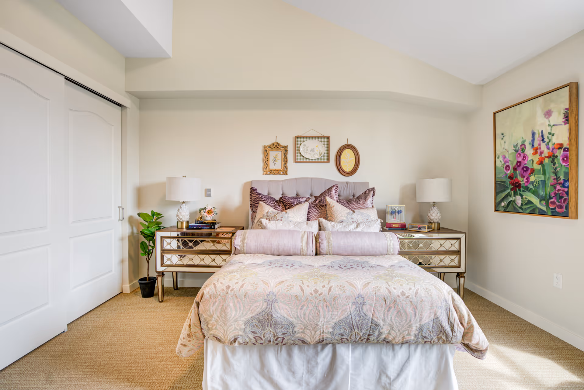 Bright furnished bedroom featuring a decorative bed with pillows and bolster, mirrored nightstands with lamps, and floral wall art.