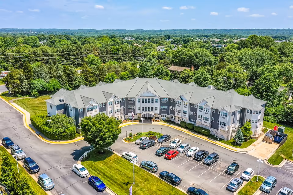 Aerial view of a curved three-story senior living building with a front circular drive, parked cars, and surrounding trees.