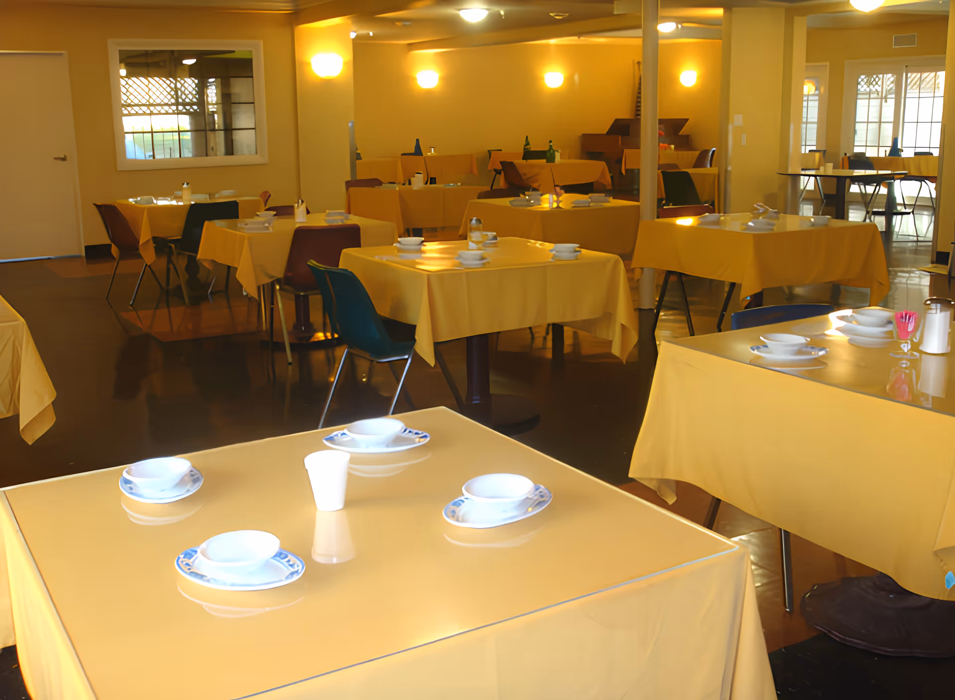 Interior view of a dining room with multiple tables covered in yellow tablecloths. Each table is set with white bowls, plates, and cups. The room is well-lit with wall-mounted lights and has a polished floor. Chairs of various colors are placed around the tables.