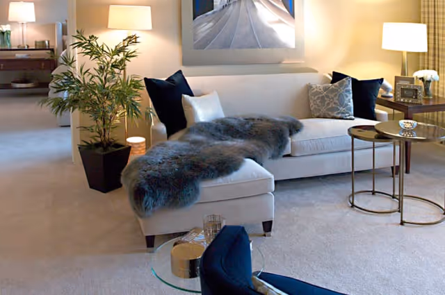 Cozy light-colored living room featuring a chaise sofa with a gray fur throw, side tables, lamps, and a potted plant.