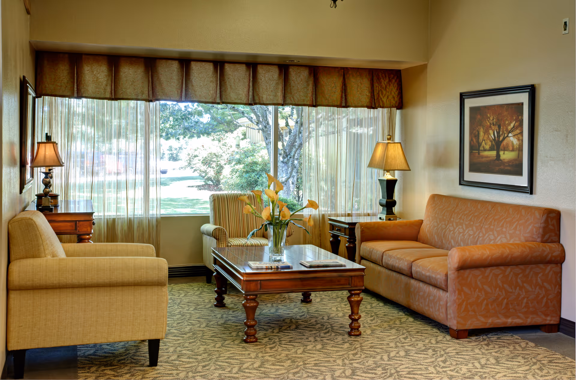 Sunlit sitting room with a sofa, armchairs and a wooden coffee table topped with flowers in front of a large window.