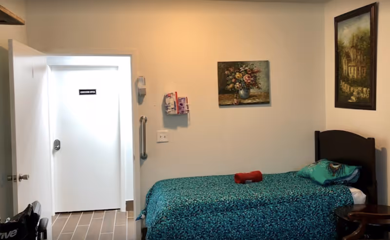 A small bedroom with a single bed covered in a blue patterned bedspread and a pillow. There is a red rolled towel on the bed. The room has two framed paintings on the walls. A door is open leading to a hallway with a closed white door labeled 'NURSING OFFICE'. A grab bar is mounted on the wall near the door.