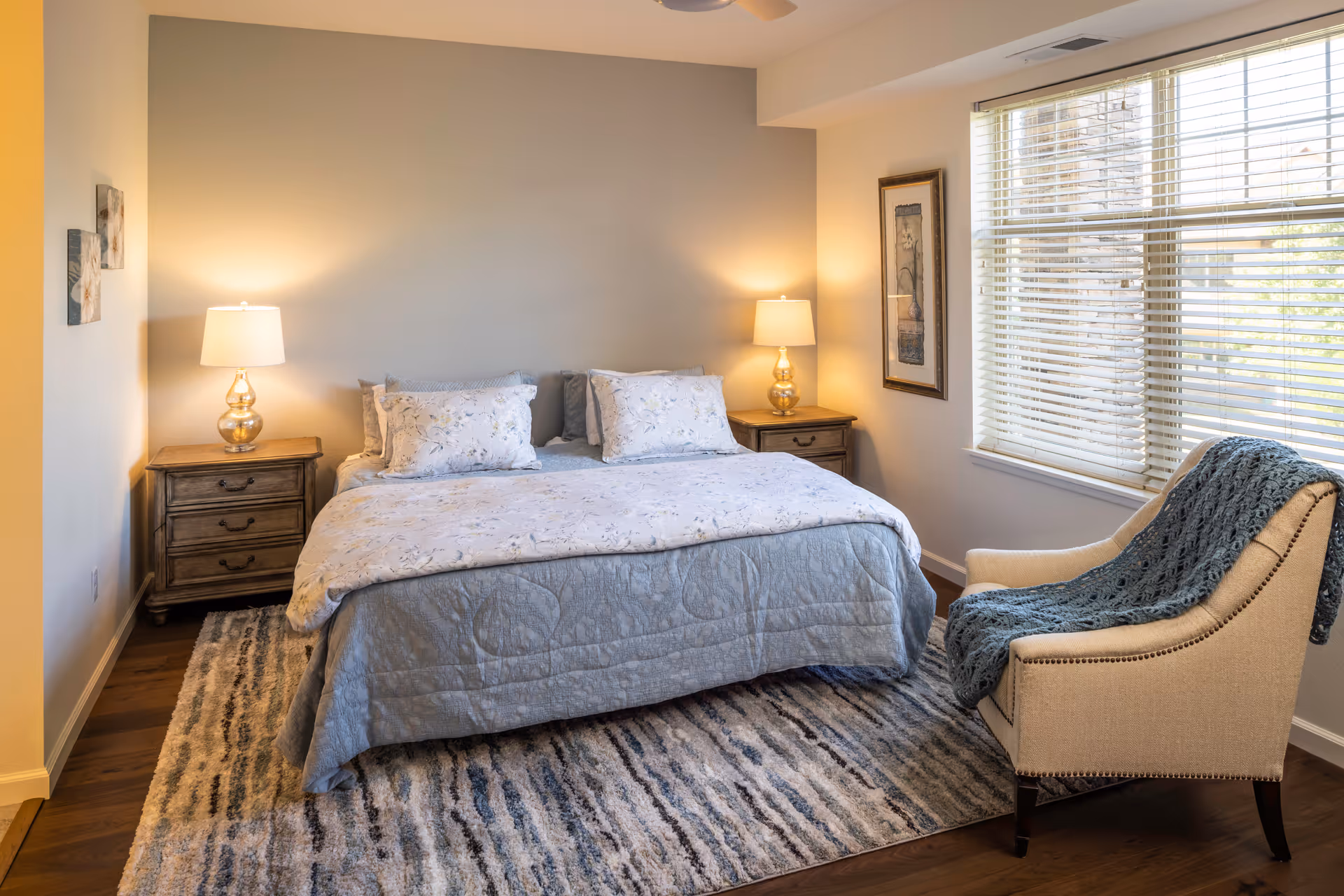 A cozy bedroom with a large bed covered in light blue and floral bedding, flanked by two wooden nightstands each with a lamp. A patterned rug lies beneath the bed, and a beige armchair with a blue throw blanket is positioned near a window with blinds. The walls are decorated with framed artwork.