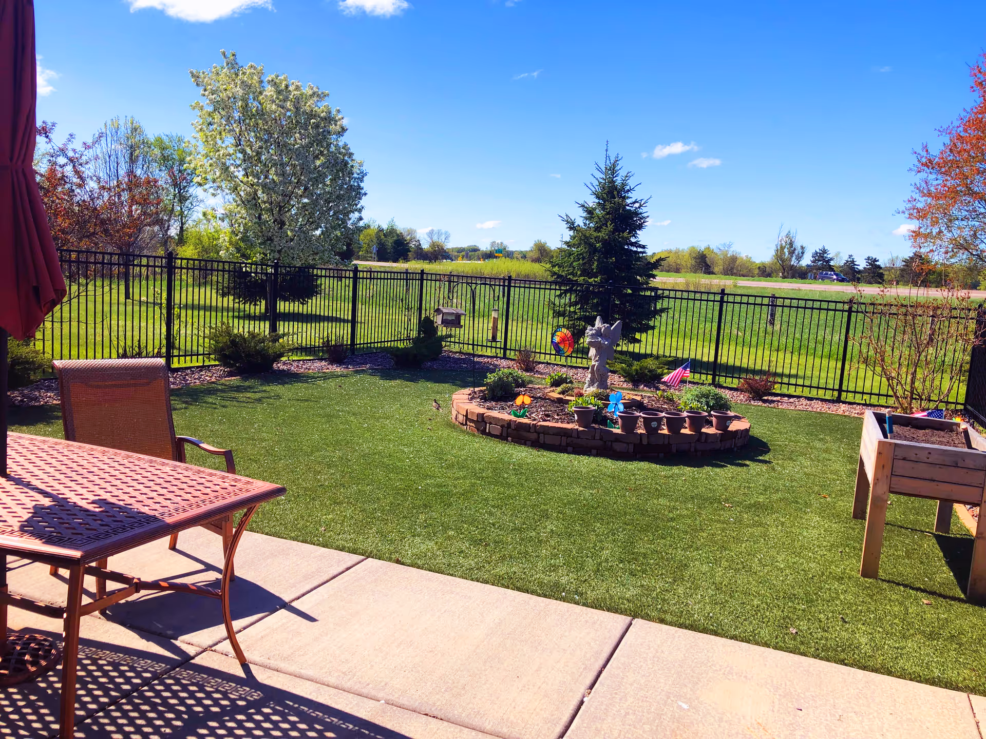 A sunny outdoor garden area with a green lawn, a circular raised flower bed with potted plants and garden decorations, a black metal fence enclosing the space, and a patio with a metal table and chair. Trees and open fields are visible in the background under a blue sky with a few clouds.