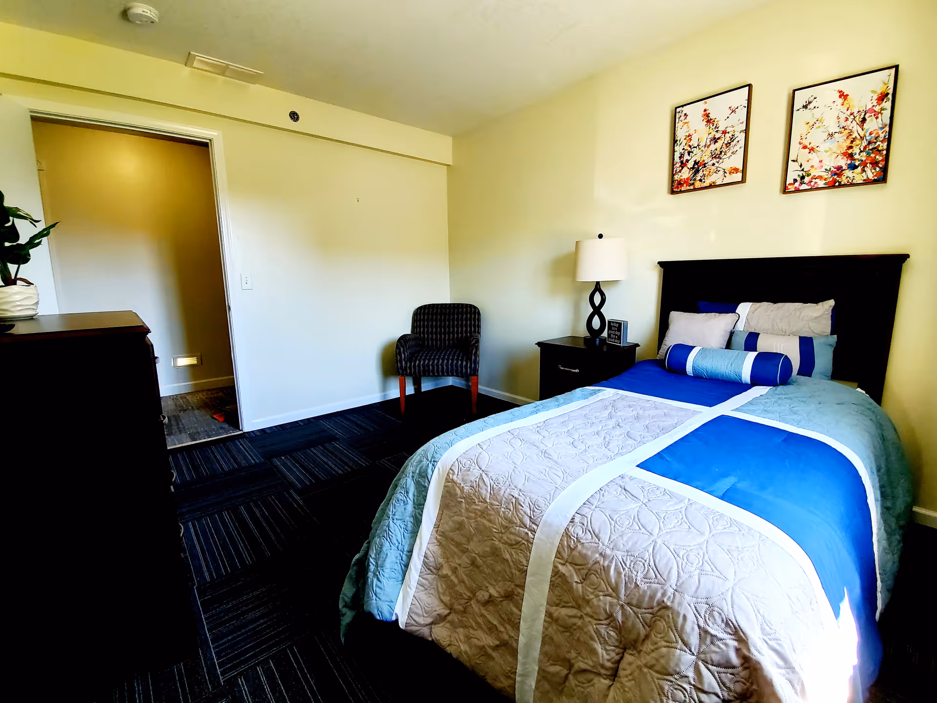 A neatly made single bed with blue and beige bedding in a senior care facility bedroom. The room has a dark wooden headboard, a nightstand with a lamp and a small decorative item, a patterned armchair, and two colorful floral paintings on the wall. The floor is carpeted with a dark pattern, and there is an open doorway leading to another area.