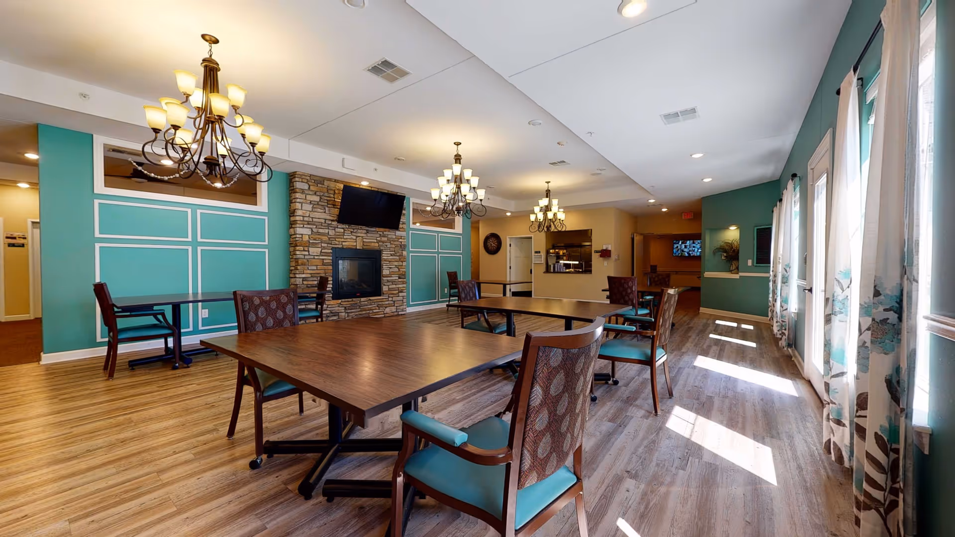 A spacious and well-lit dining room in a retirement living facility featuring wooden tables and cushioned chairs with patterned backs. The room has teal accent walls with white trim, a stone fireplace with a mounted TV above it, and multiple chandeliers hanging from the ceiling. Large windows with floral curtains allow natural light to fill the space, and there is a kitchen area visible in the background.