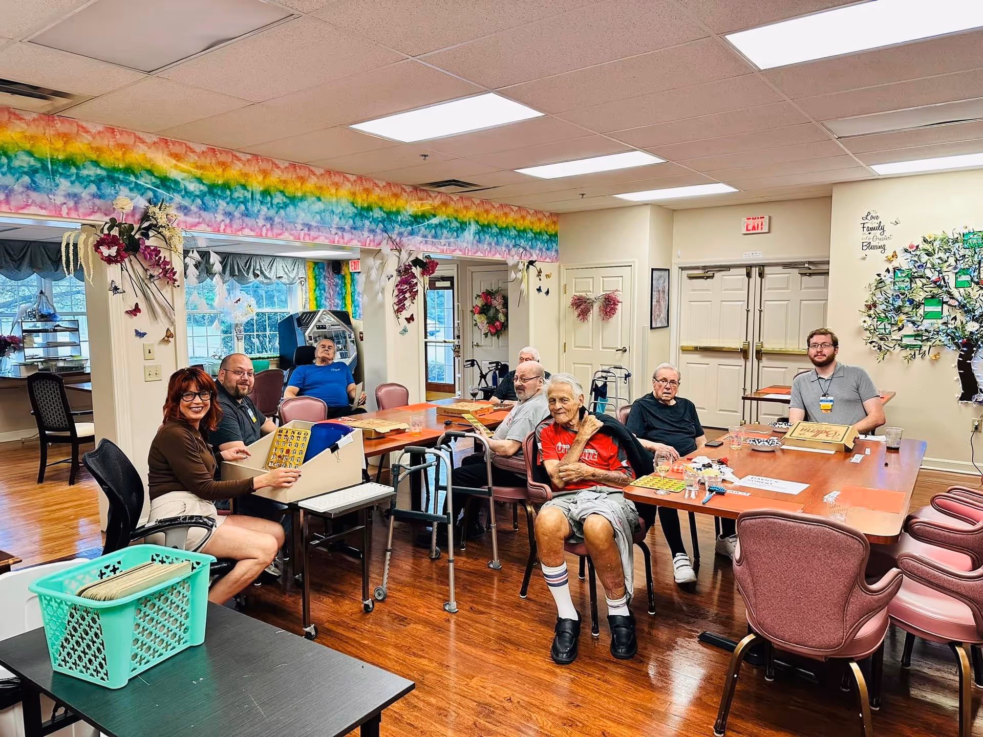 A group of elderly people and two staff members sitting around tables in a brightly decorated common room with rainbow-colored wall decorations, playing bingo and socializing. The room has wooden floors, large windows, and a wall decorated with a tree and butterflies.