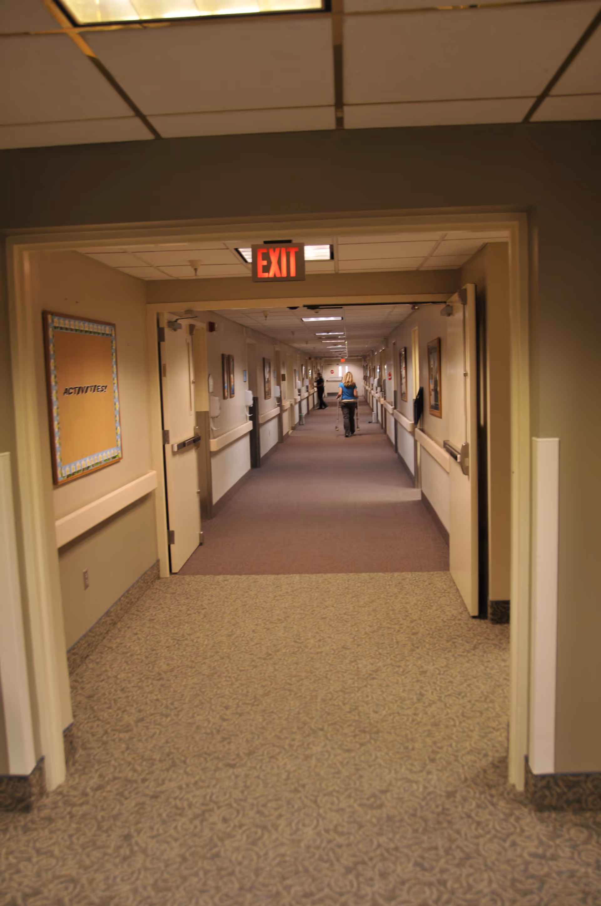 A long hallway in a skilled nursing and rehabilitation center with beige walls and carpeted floors. There are handrails along both sides of the hallway and several closed doors. A person is seen walking away in the distance. An exit sign is visible above the doorway in the foreground, and a bulletin board labeled 'ACTIVITIES' is on the left wall.