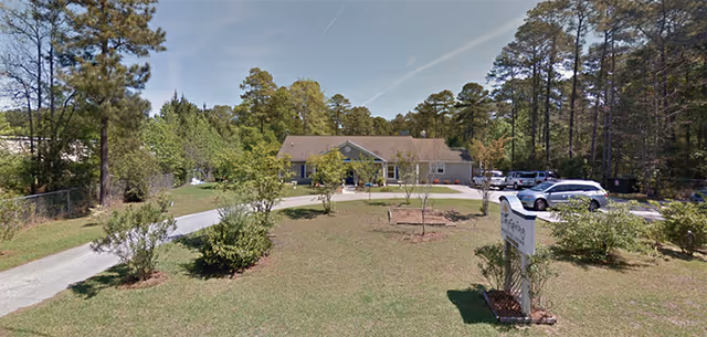 Exterior view of Dayspring Assisted Living facility showing a single-story building surrounded by trees and greenery. There is a driveway leading to the entrance with several parked cars and a sign with the facility's name in the foreground.