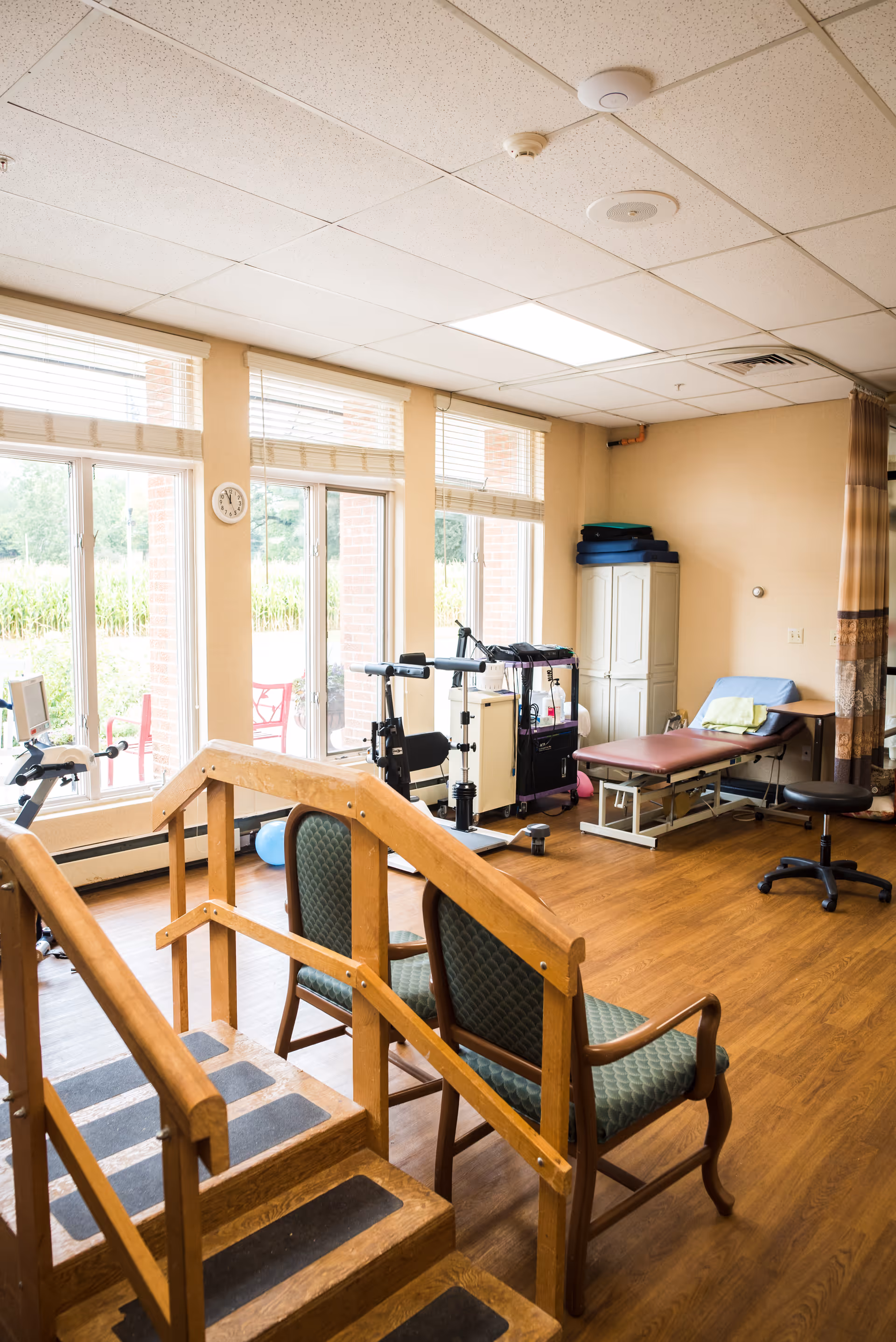 A rehabilitation room with large windows letting in natural light. The room contains a wooden staircase with handrails for physical therapy, two green cushioned chairs, exercise equipment, a treatment table with a pillow and towel, and a rolling stool. The floor is wooden, and the walls are beige.