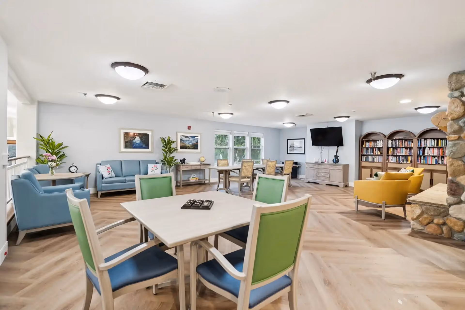 A bright and spacious common area in a senior living facility featuring multiple seating arrangements including blue and yellow armchairs, green and blue chairs around a square table, a blue sofa, and a wooden table with chairs near windows. The room has light wood flooring, a stone fireplace, bookshelves filled with books, framed pictures on the walls, and a wall-mounted TV.
