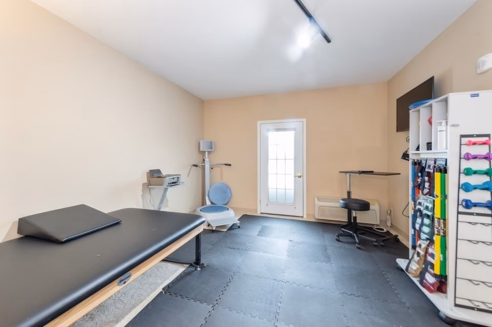 Small therapy/exercise room with a treatment table, exercise equipment, weights, and a door.