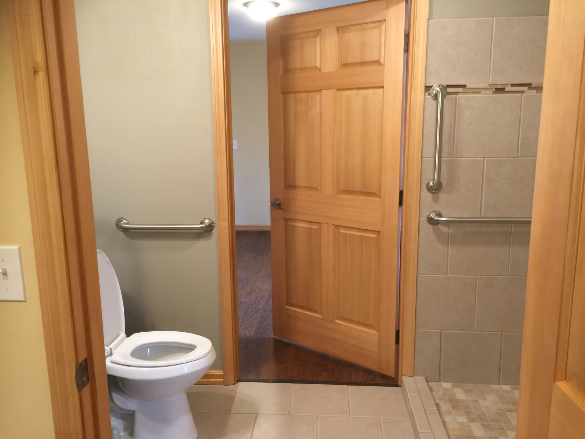 Interior view of a bathroom with a white toilet on the left and a tiled walk-in shower with grab bars on the right. The bathroom has beige walls and light-colored floor tiles. A wooden door is open, showing a glimpse of a room with wooden flooring outside the bathroom.