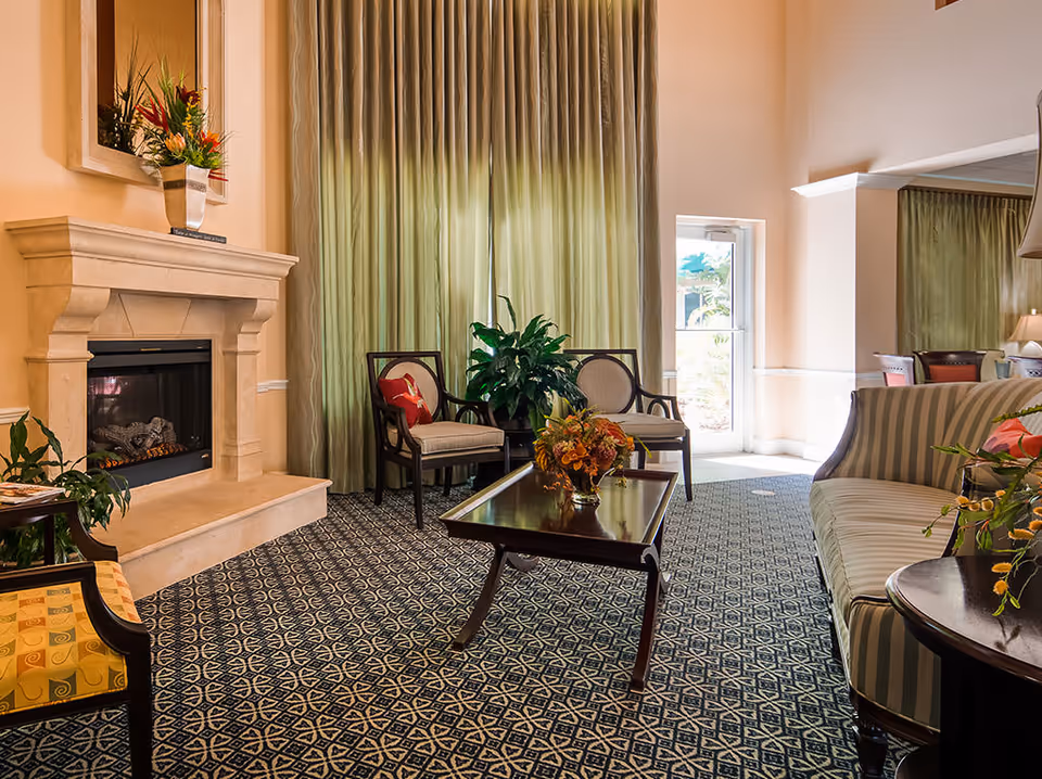 A bright sitting area with a fireplace, upholstered chairs and sofa, a coffee table with flowers, and tall green curtains.
