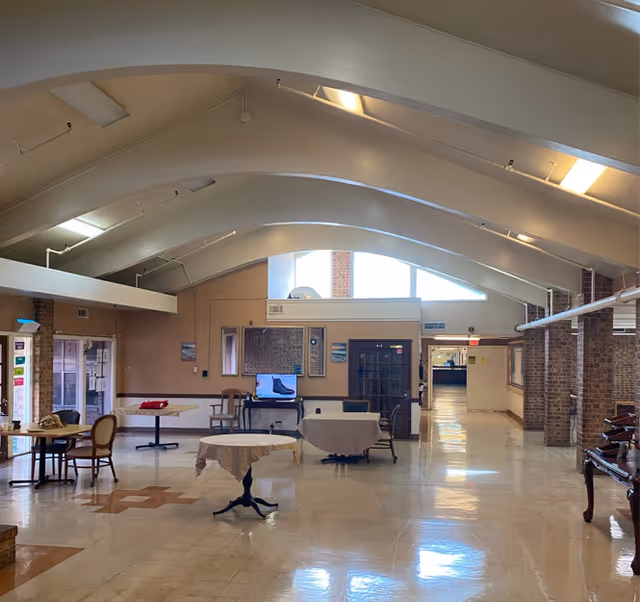A spacious interior common area with high arched ceilings and large windows letting in natural light. The room has several round tables with chairs around them, a television on a stand, and a polished floor. The walls are a mix of brick and light-colored paint, and there is a hallway leading further into the building.