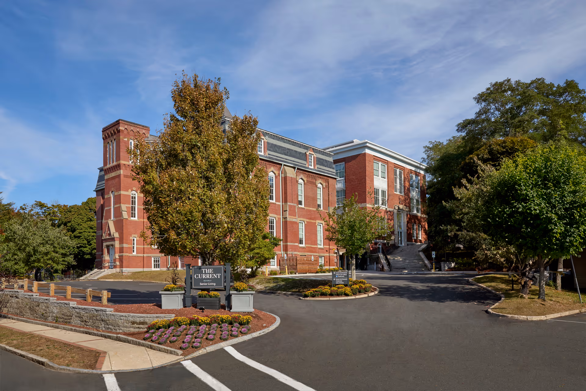 Exterior view of a large red brick building with multiple windows and a tower-like structure on the left side. The building is surrounded by trees and landscaping, with a driveway and parking area in front. A sign near the entrance reads 'The Current Senior Living'. The sky is clear with some clouds.