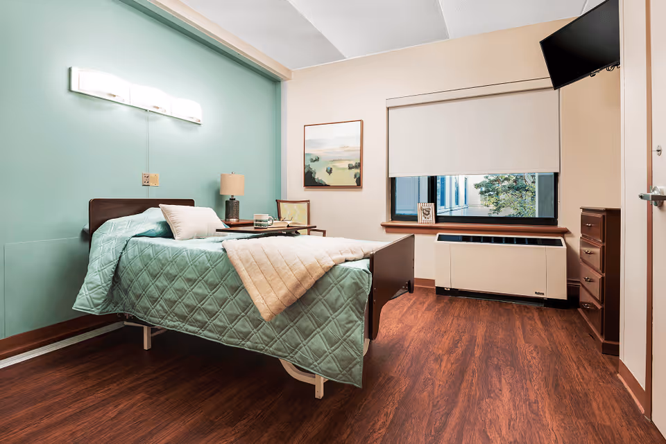 A neatly arranged bedroom in a senior living facility with a single bed covered in a light green quilt and a beige blanket. The room features a wooden floor, a bedside table with a lamp and a cup, a chair, a framed landscape painting on the wall, a window with a white roller blind partially down, a wall-mounted TV, and a heating/cooling unit beneath the window.