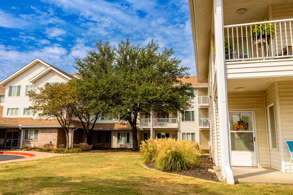 Exterior view of a multi-story senior living facility building with beige siding and brick accents, surrounded by green grass, trees, and ornamental grasses under a partly cloudy blue sky.