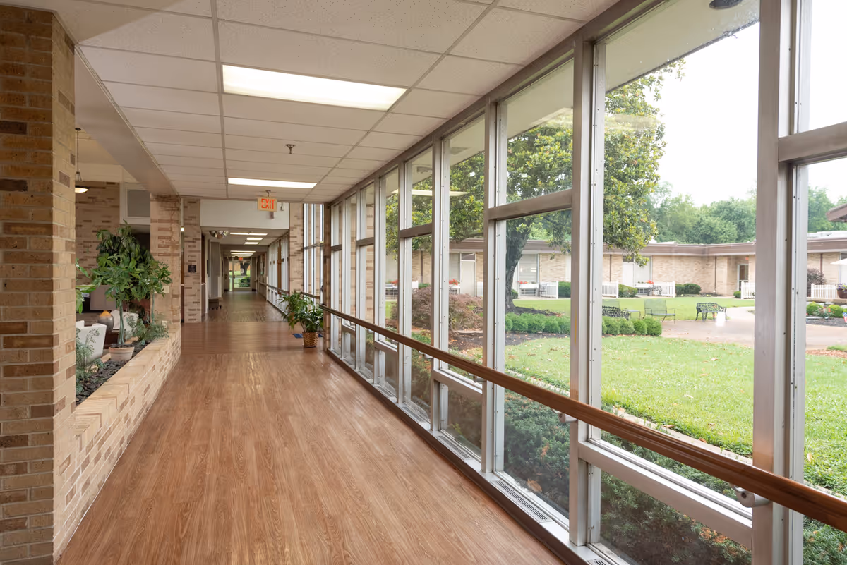 Long indoor hallway with large floor-to-ceiling windows and wooden handrails overlooking a landscaped courtyard.