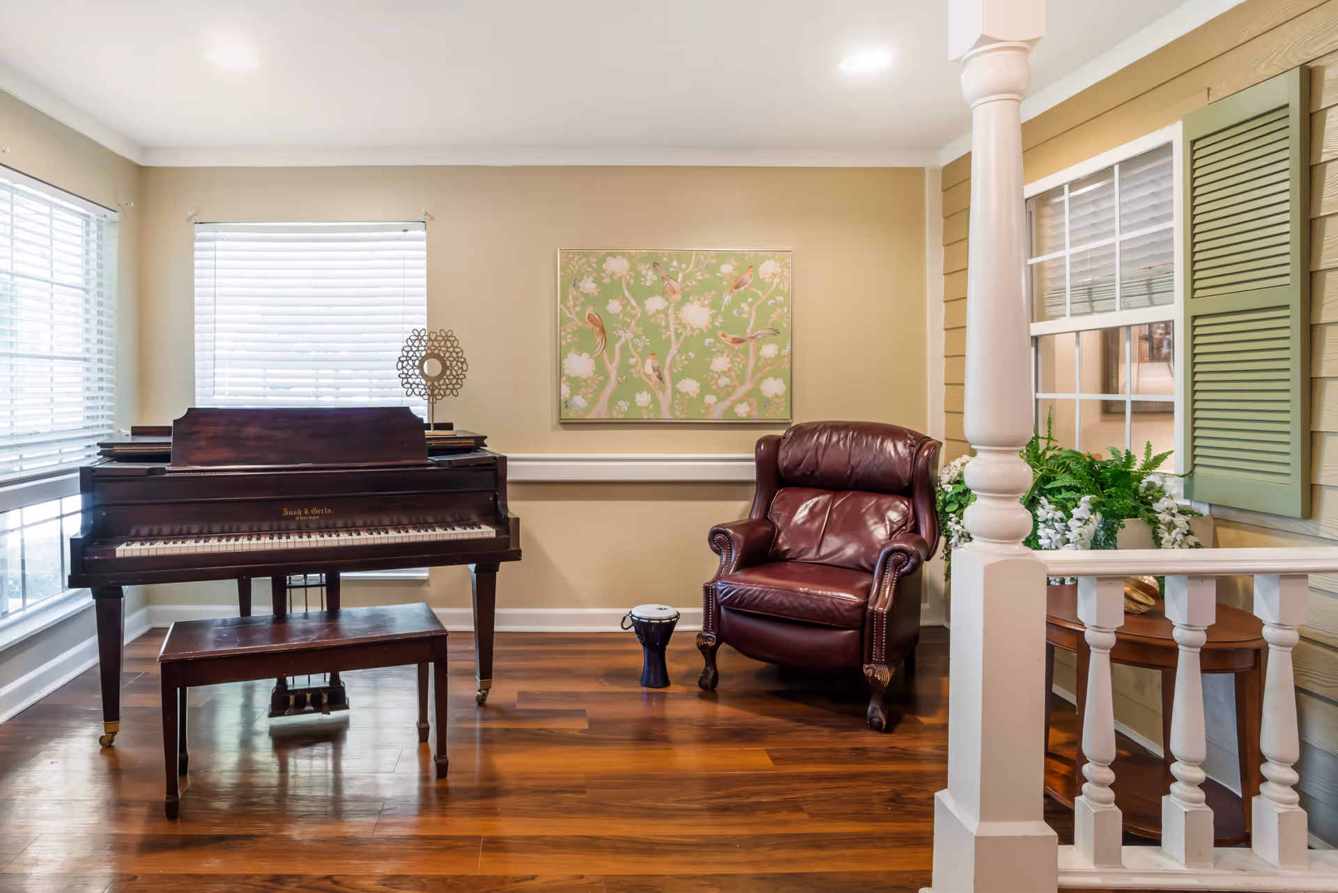 A cozy interior room featuring a dark wooden piano with a matching bench, a burgundy leather armchair, a small drum on the wooden floor, and a decorative floral painting on the beige wall. The room has large windows with white blinds and a white column with railing in the foreground, along with a window with green shutters and a table with green plants and white flowers.