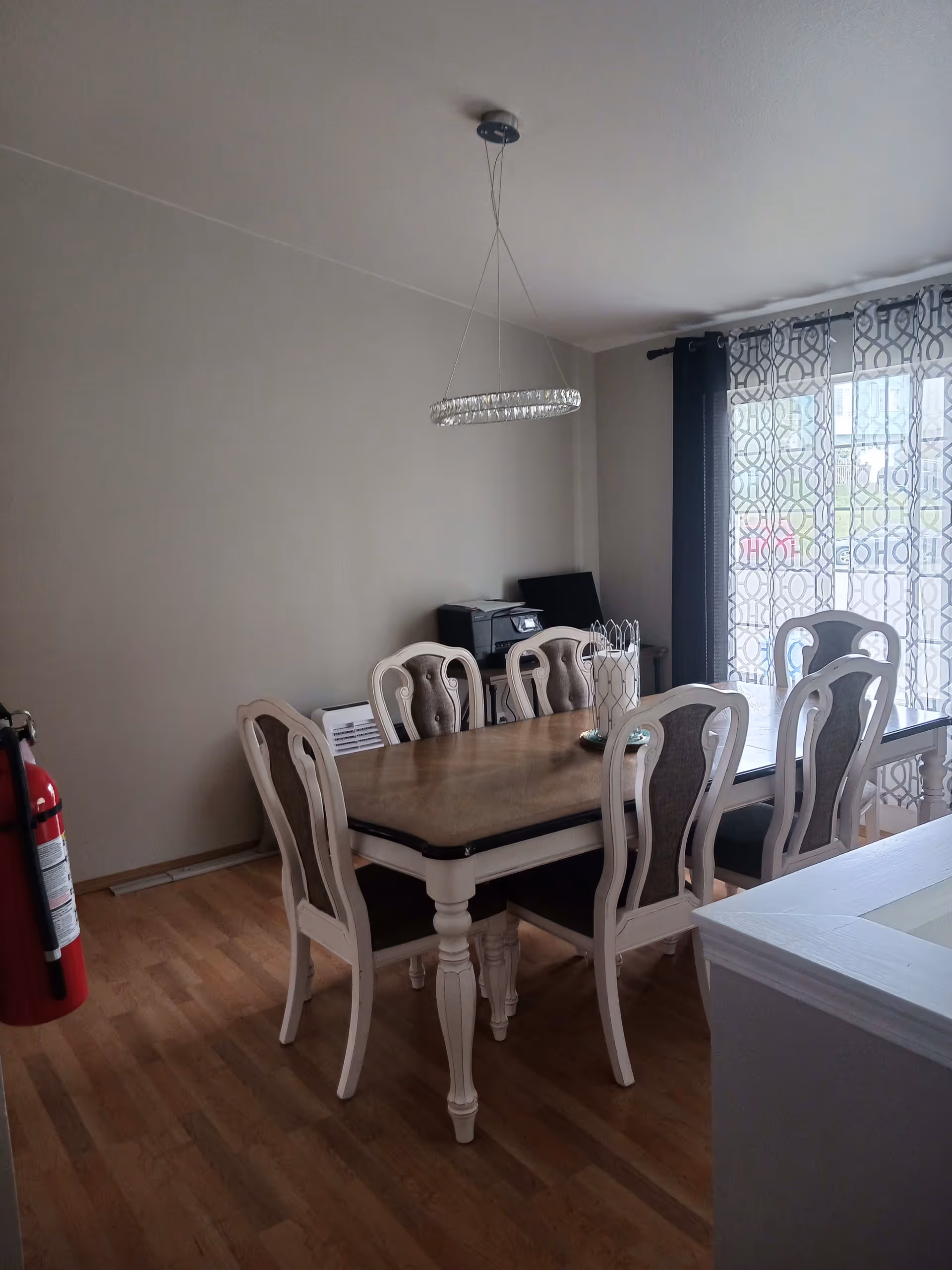 A dining room with a wooden dining table and six matching chairs with cushioned backs and seats. A modern circular chandelier hangs above the table. There is a window with patterned curtains allowing natural light into the room. A fire extinguisher is mounted on the wall to the left, and a printer is placed on a small table in the corner.
